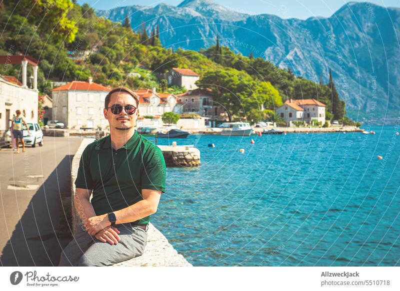 Middle-aged man with green shirt and sunglasses sits on a wall on the coast of Perast in Montenegro Mediterranean sea Water Vacation & Travel Idyll