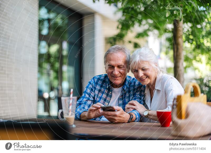 Senior couple sitting at a table in a cafe and using mobile phone people standing healthy city life gray hair enjoy street casual day portrait outside