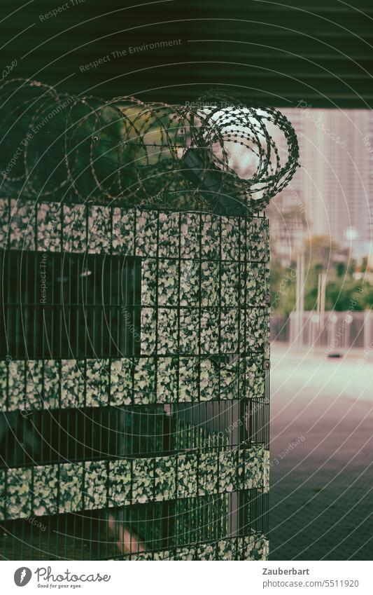 Cityscape in movie look, construction fence with barbed wire and green fake foliage, behind it concrete pillars of a bridge Town Landscape Alarming Hoarding