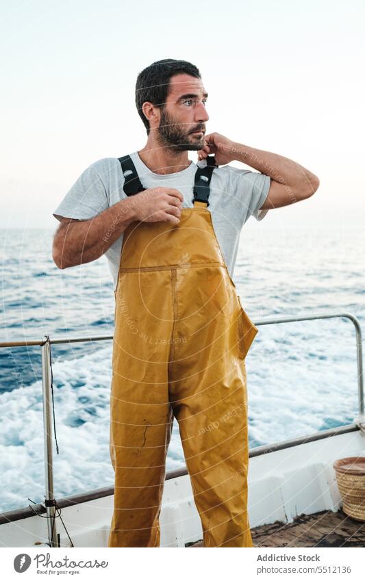Confident young ethnic fisherman tightening braces of bib on sailboat travel fasten waterproof focus trip male hispanic concentrate journey adventure transport