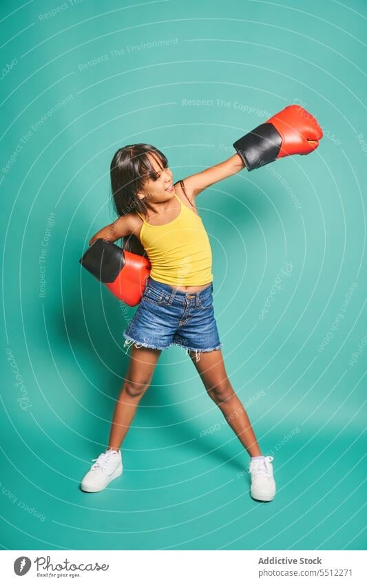 Cheerful ethnic kid girl in boxing gloves throwing punches against turquoise backdrop child smile childhood having fun happy playful asian sport cheerful