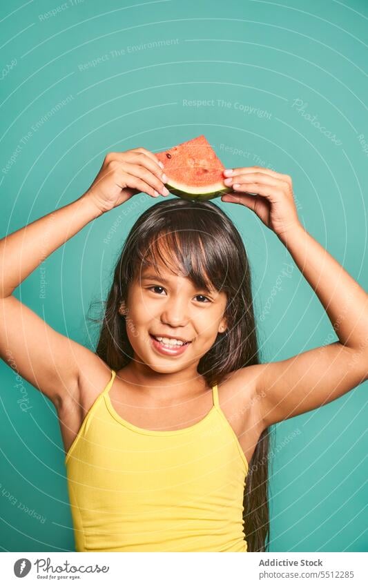 Happy ethnic girl child standing with piece of watermelon against turquoise backdrop kid portrait childhood show cute delicious happy playful asian black hair