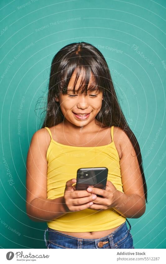 Funny ethnic girl child standing with smartphone near turquoise backdrop kid funny using cute smile cheerful asian mobile device gadget adorable happy preschool