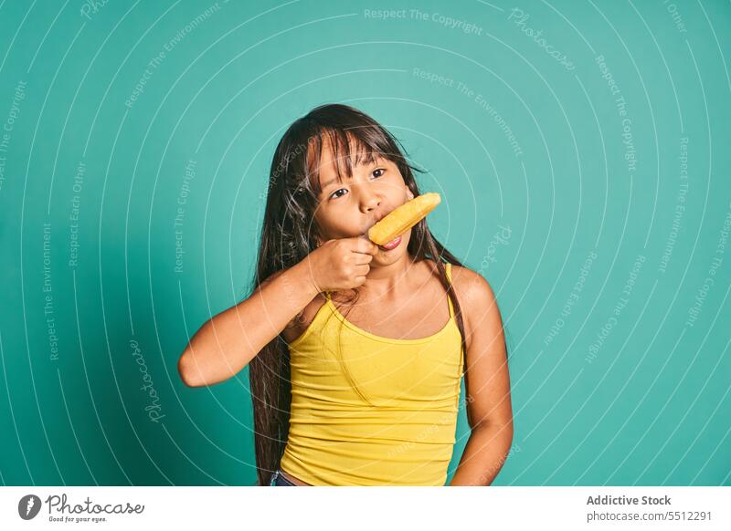 Happy ethnic girl child standing and eating ice cream in hand against turquoise backdrop kid popsicle delicious appearance sweet cute tasty adorable asian