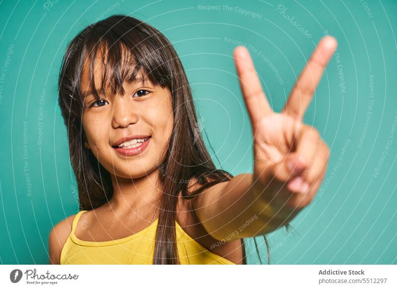 Happy ethnic girl child standing and showing victory sign against turquoise backdrop gesture smile kid v sign two fingers happy cute positive joy asian