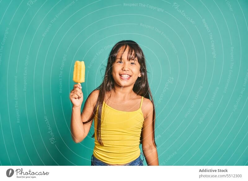 Happy ethnic girl child standing with popsicle in hand against turquoise backdrop kid childhood show frozen cute delicious happy playful asian black hair