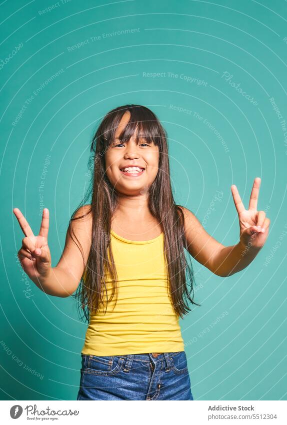 Happy ethnic girl child standing and showing victory sign against turquoise backdrop gesture smile kid v sign two fingers happy cute positive joy asian