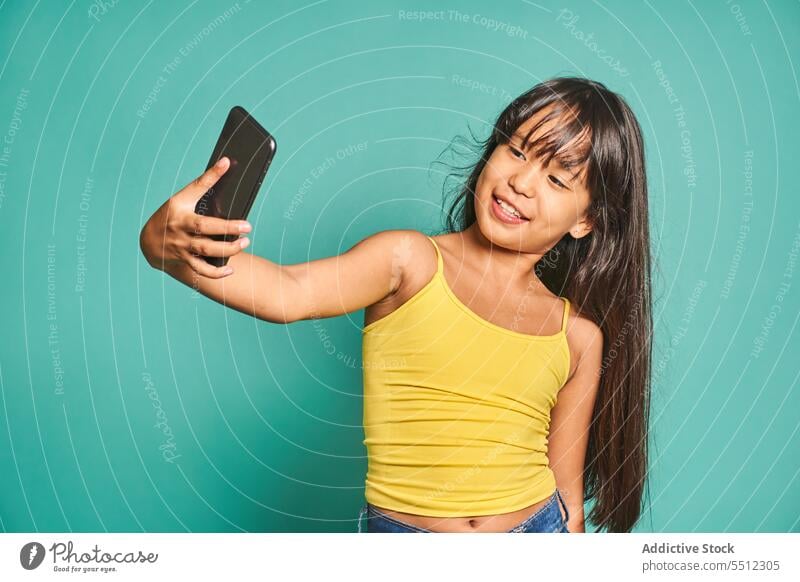 Funny ethnic girl child standing with smartphone and taking selfie near turquoise backdrop kid funny using cute smile cheerful asian mobile device gadget