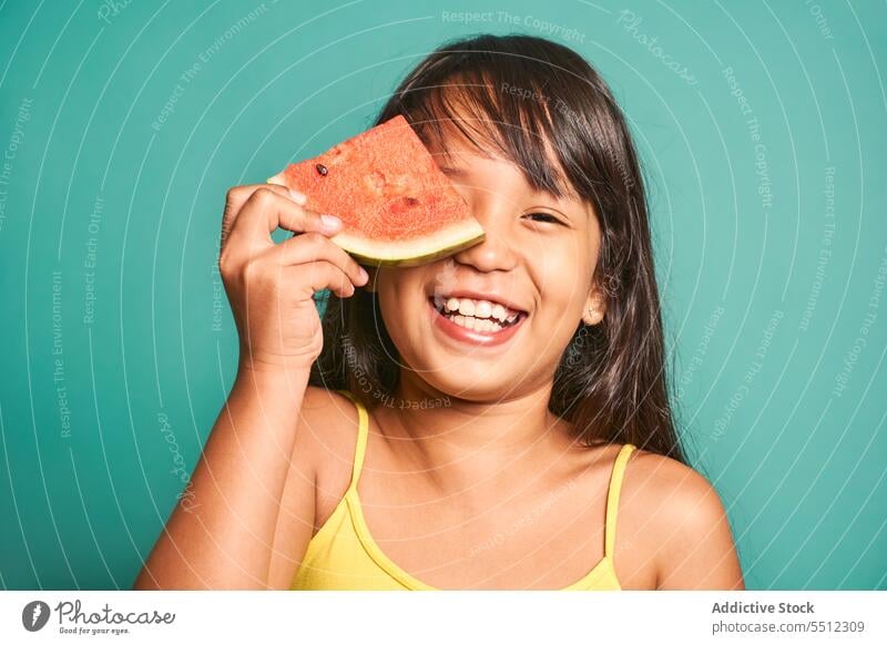 Happy ethnic girl child standing piece of watermelon near face against turquoise backdrop sweet cute delicious smile adorable kid asian colorful tasty treat