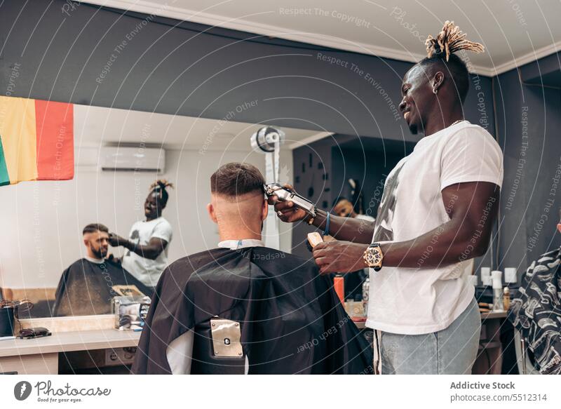 Happy barber trimming hair of male client men haircut trimmer barbershop accuracy beauty young multiracial african american black ethnic cape machine salon