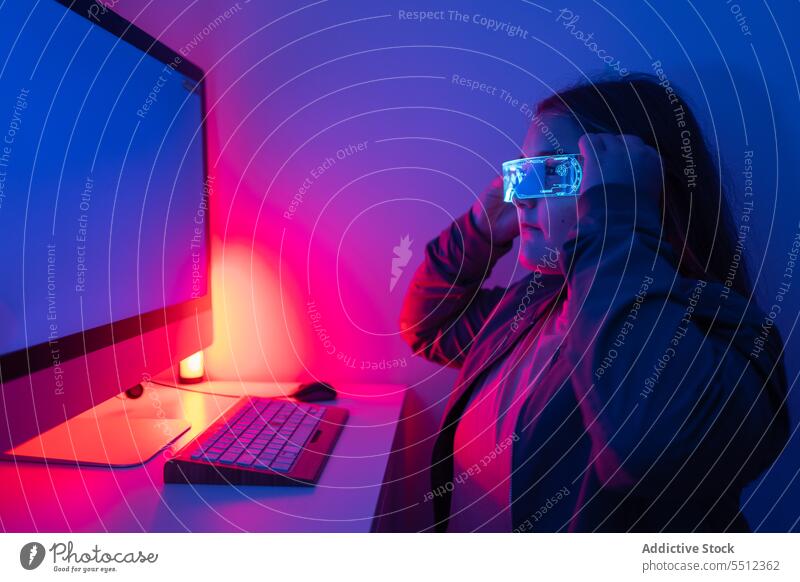 Young girl wearing AR glasses sitting at desk with computer monitor teenage screen neon table corner room gadget device light display online serious illuminate