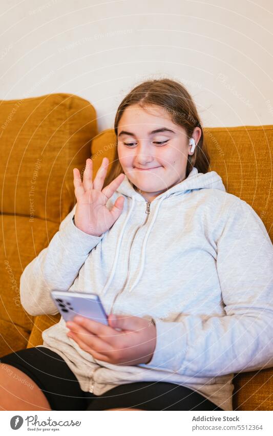 Cheerful young girl saying hi at smartphone while sitting on sofa video call smile hello online using communicate connection internet teen gadget device chat