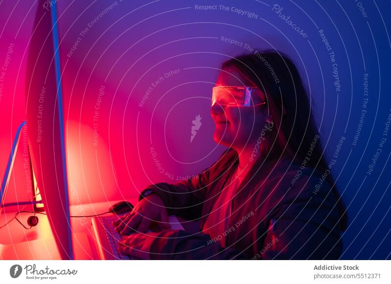 Young girl wearing AR glasses sitting at desk with computer monitor teenage screen neon table smile corner room gadget device light display online illuminate