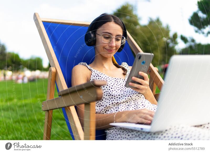 Smiling woman with laptop and smartphone on deckchair using park positive mobile summer browsing female smile young internet surfing happy device gadget