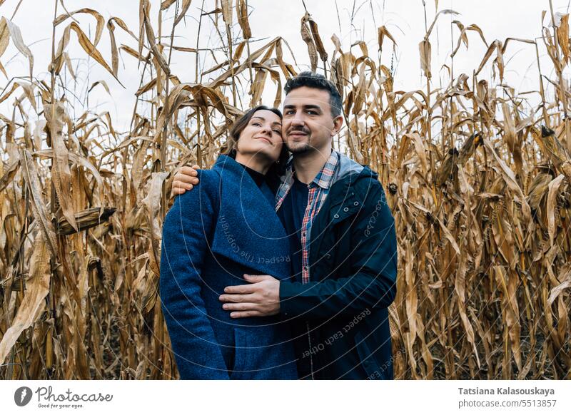 A young man hugs a woman standing against the backdrop of a cornfield in autumn couple embrace love affection relationship rural coat jacket countryside