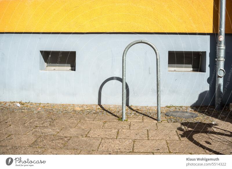 Light and shadow on a house wall. A bicycle stand made of galvanized steel pipe and a rain gutter just like it stand in front of the yellow/blue facade. The shadow of a bicycle pushes into the picture from the right.