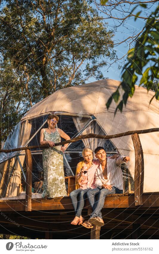Smiling couple with standing mature woman sitting on wooden structure with camp tent happy glamping smile countryside show summer male female young sunglasses