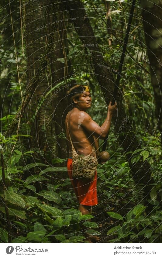 Tribal man with long stick in forest shirtless tropical nature bag environment serious exotic green male summer woods plant tree foliage natural flora season