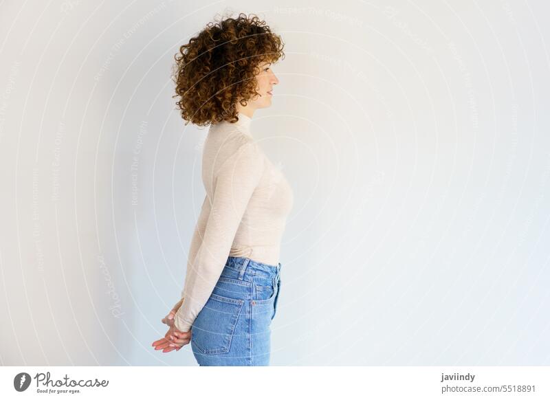 Curly haired woman standing with hands behind back hand behind back hands clasped studio shot straight curly hair posture jeans turtleneck unemotional