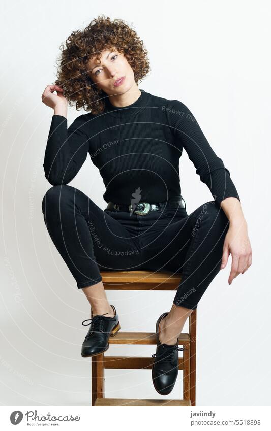 Stylish woman in black dress sitting on wooden bench stool touch hair fashion step tilt leg raised curly hair relax model apparel female appearance feminine