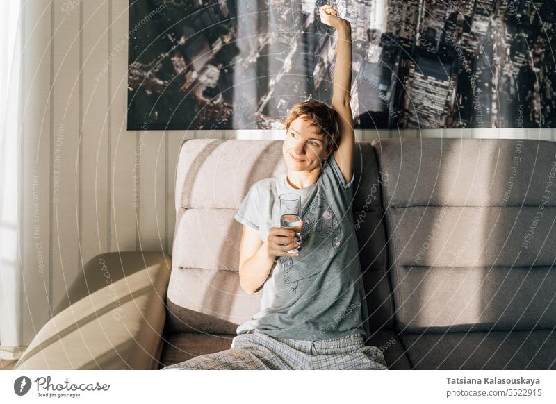 A young woman stretches, smiles and holds a glass of water sitting on the couch Lifestyles female people happiness Drink person Drinking caucasian Glass