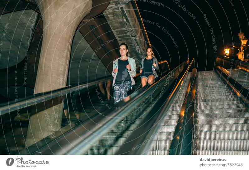 Women friends training running down escalator in city at night women female runner mechanic stair staircase step light leg action speed copy space happy