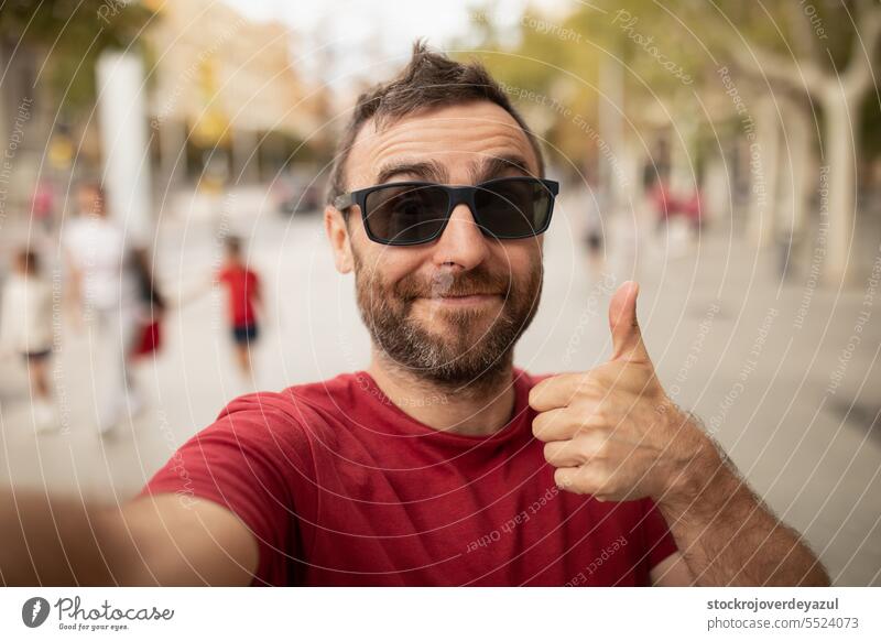 A young, bearded man, wearing sunglasses, smiles at the camera, while taking a selfie with his cell phone during a walk in the city. person men happy thumbs up