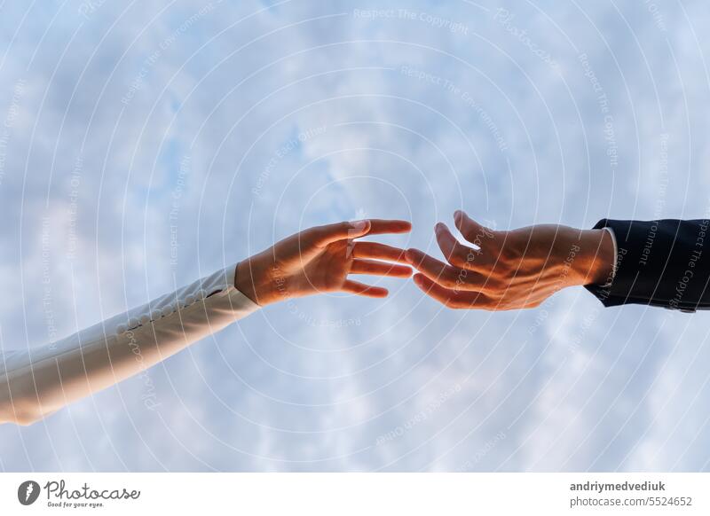 Hands of bride in white dress and groom in suit reaching each other, touching fingers on blue sky background. Helping hands for save and support people concept. Wedding day. Valentine day.