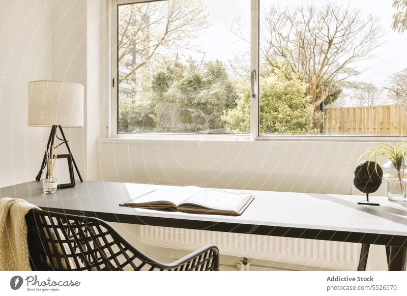Home office with book and lamp on desk against window small plant chair glass large bright light sunlight education white wall copy space house apartment study