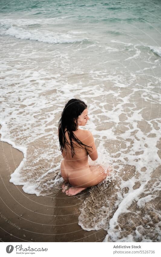 Naked woman relaxing in seawater on shore nude sensual beach seashore foam summer vacation wet hair sand coast female kneel ocean rest slim naked recreation