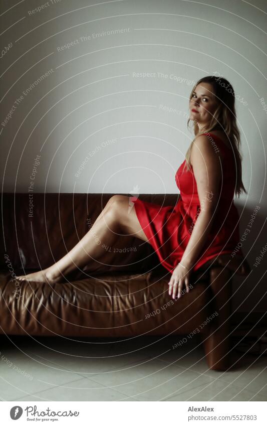 Blonde woman in red dress sits comfortably on brown leather couch Woman portrait Long-haired long hairs Face naturally pretty Authentic Attractive