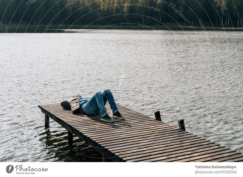 A man relaxes lying on a wooden boat dock on a lake in autumn Man jeans jumper jacket lies relaxing enjoying sunny day resting outside pier rests adult