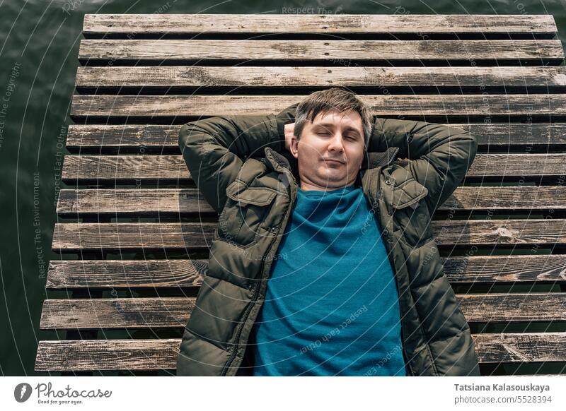 Top view of a man lying on a wooden boardwalk boat dock on a lake with his eyes closed in a jumper and jacket Man lies relaxing enjoying sunny autumn day