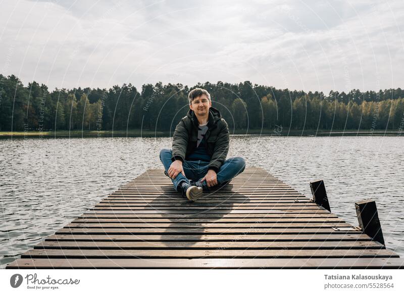A man enjoys a sunny autumn day while sitting on a wooden pier on a lake jeans jumper jacket relaxing boat dock enjoying relaxes resting sits cross-legged