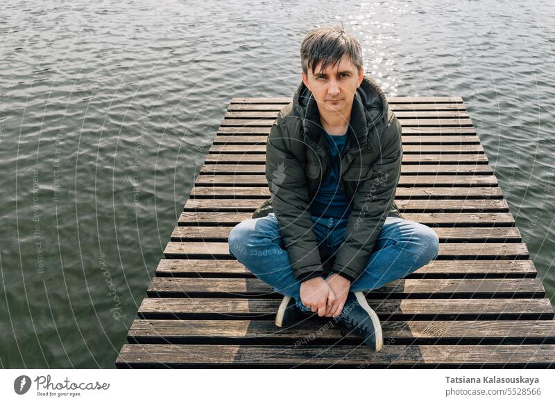 An adult man sits cross-legged on a wooden pier on a lake in jeans and a jacket jumper relaxing boat dock enjoying sunny autumn day relaxes sitting resting