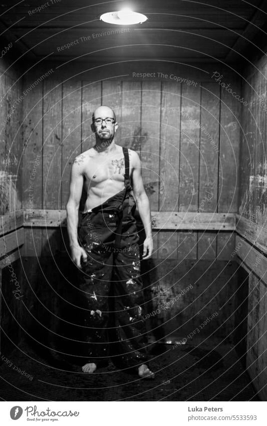 A muscular middle-aged man with a beard and bald head in partially opened, paint-stained dungarees and a naked torso stands in an old elevator and looks directly into the camera.