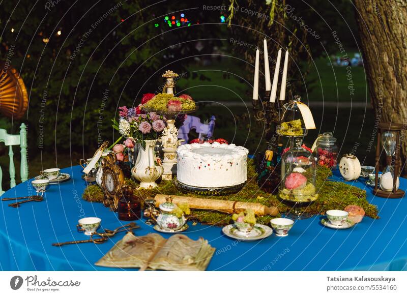 The dining table is decorated in the style of Alice in Wonderland. An open old book, a cake, tea bowls, candles in a candlestick, an hourglass on a blue tablecloth late in the evening