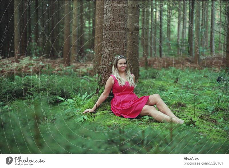 analog portrait of beautiful blonde long haired woman sitting in a light forest leaning against a tree and looking into the camera Emotions Attractive long legs