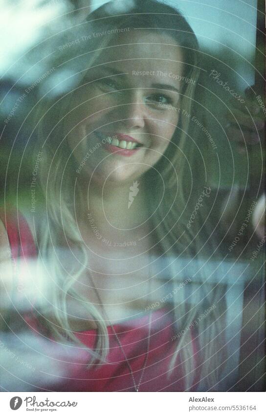 Blonde woman looks through a window pane with reflections directly into the camera and smiles - analog portrait Woman Long-haired long hairs Reflection Face