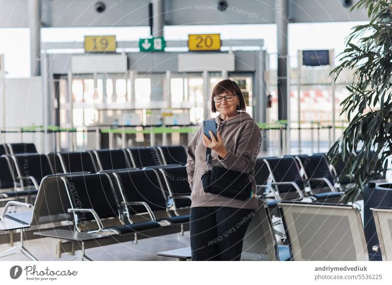Happy smiling adult traveler woman holds smartphone, has video call, waves hand in greeting. Aged vlogger or blogger has live at airport terminal waiting for flight, social media. Holiday, vacation