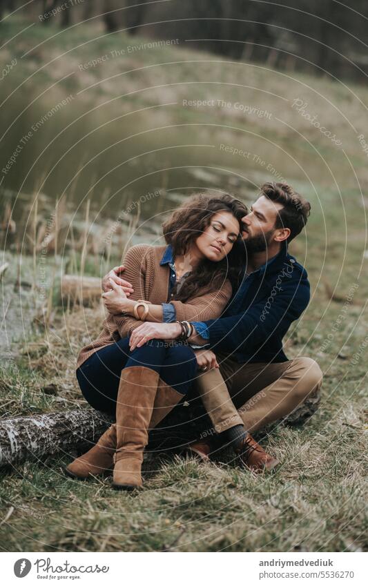 Happy lovers are sitting on the shore of the lake. Young couple is hugging on autumn day outdoors. A bearded man and curly woman in love. Valentine's Day. Concept of love and family.