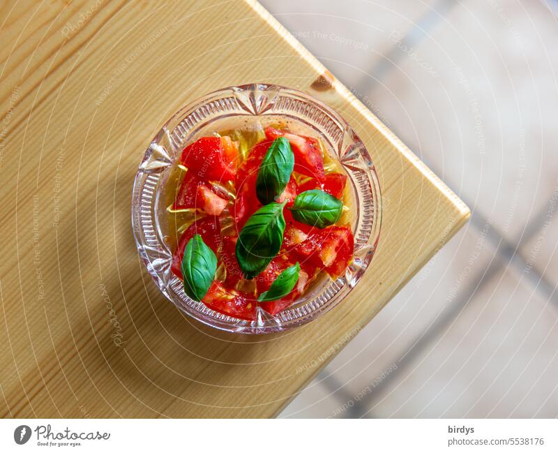 Fresh tomatoes with olive oil and fresh basil in a glass bowl Tomato salad Basil Basil leaves Glass bowl Table edge Bird's-eye view Wooden table Delicious