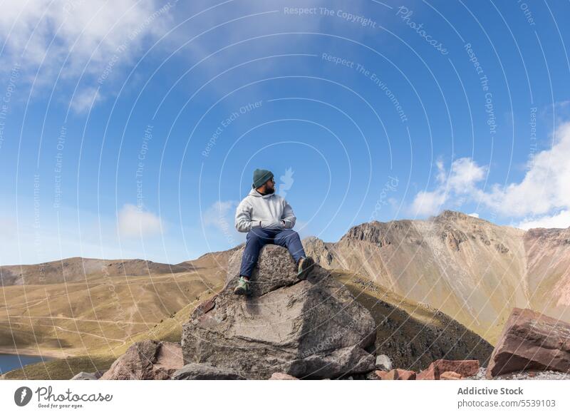 Young man in cap warm clothes sitting on boulder on mountain traveler rock volcano landscape admire explore nature male young hand in pocket cloudy journey