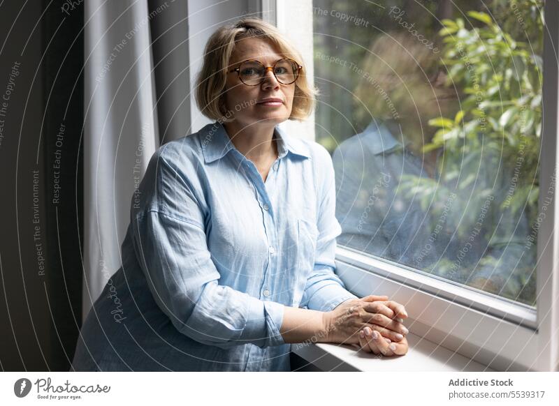 Mature blond woman near the window at home care room apartment light tranquil daylight living room indoors blonde mature female tree reflection alone eyeglasses
