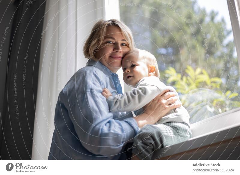 Mature blond woman and a baby boy near the window at home mother infant child love care childcare parent motherhood childhood family room parenthood bonding