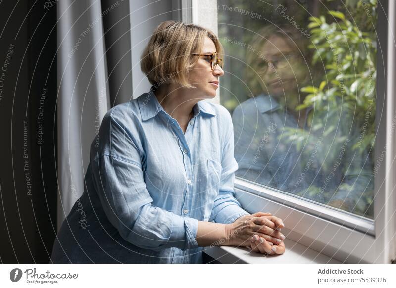 Mature blond woman near the window at home care room apartment light tranquil daylight living room indoors blonde mature female tree reflection side view alone