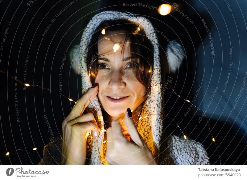 Portrait of a young woman in a room in a hood decorated with a fairy lights string Illuminated Tangled Night Dark Head Evening electric christmas lights