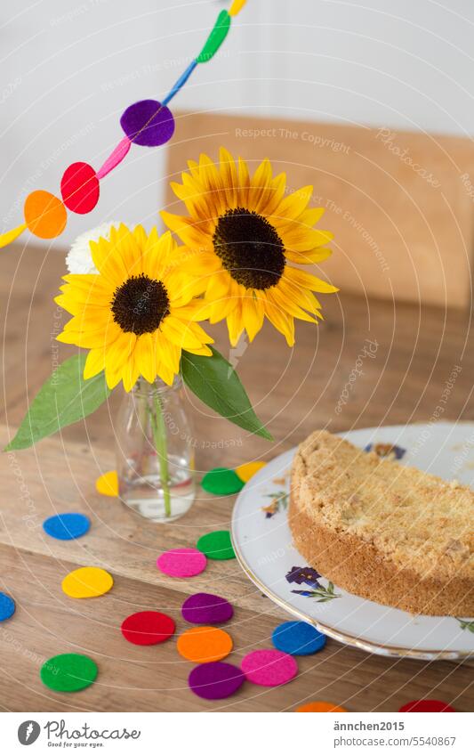 on a wooden table are sunflowers in a small vase next to it is a cake and you can see confetti and a colorful garland Sunflowers Ostrich Confetti celebrations