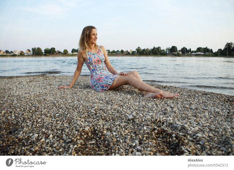 A beautiful, slim, young woman in a summer dress sits barefoot on a sandbank of gravel in the Rhine and looks into the sunset Woman beautiful woman Slim