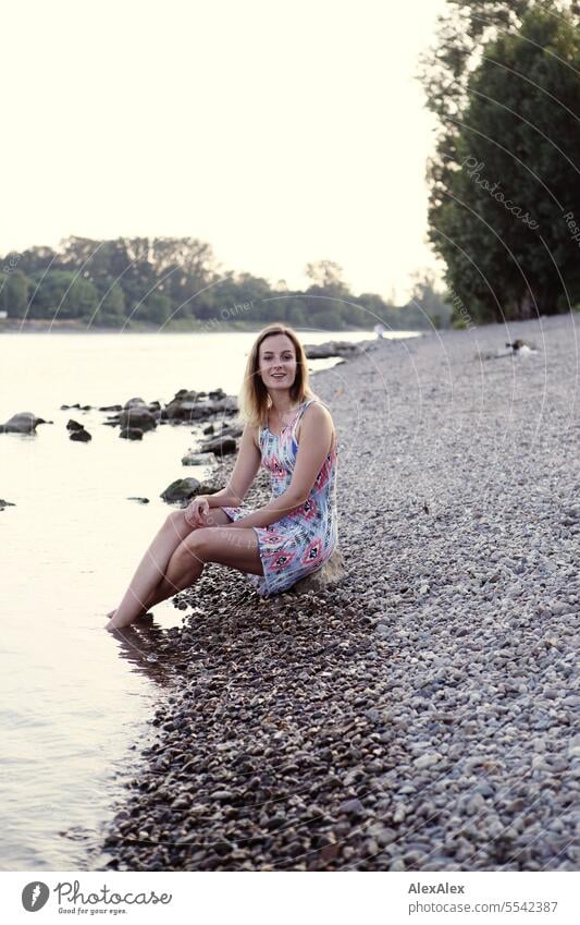 Beautiful slim young woman in summer dress sits barefoot on Rhine shore made of gravel with feet in water Woman beautiful woman Slim Athletic youthful teen